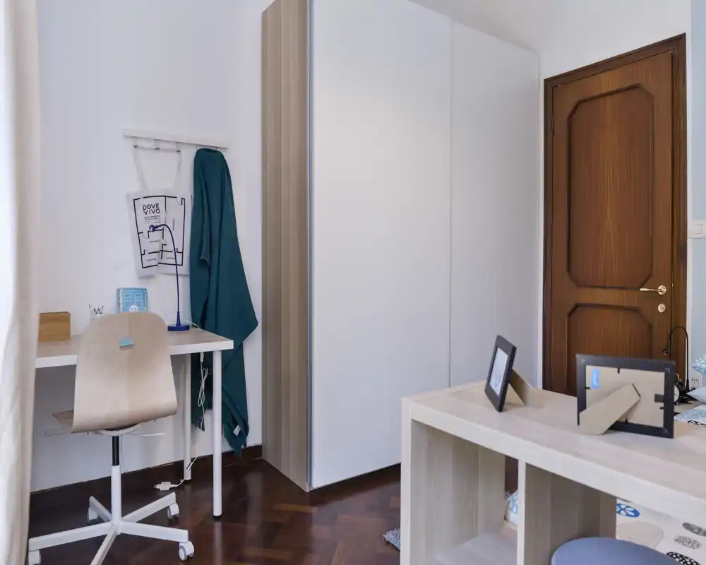 Compact bedroom corner with a study desk, wardrobe and shelving; bright, tidy space ideal for a single occupant or student.
