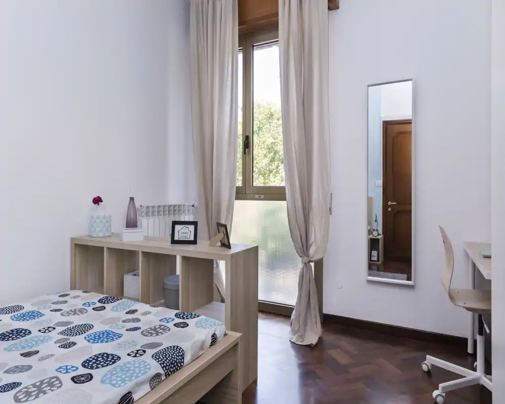 Bright, tidy bedroom with a patterned double bed, light wood furniture and full-length mirror. Natural light from the window and neutral curtains create a calm, airy atmosphere ideal for relaxing stays.