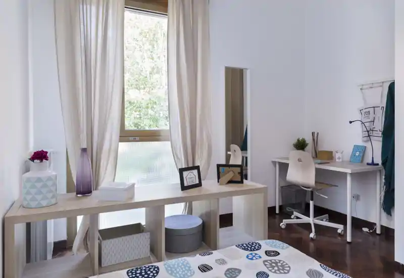 Bright, modern bedroom with patterned bedcover, light wood furniture and a small workspace by the window — cozy and well-lit for relaxing or working.
