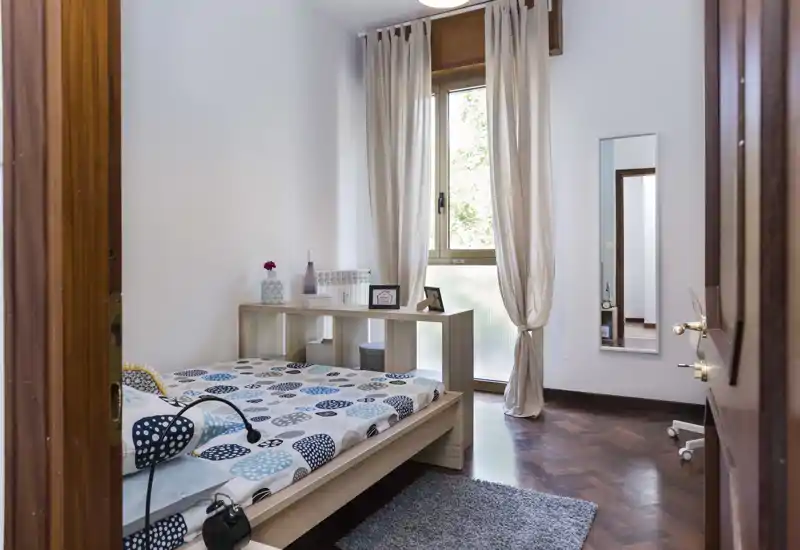 Bright, neatly styled single bedroom with a wooden bed frame, patterned bedding, a small shelving desk at the foot of the bed and a large window letting in natural light.