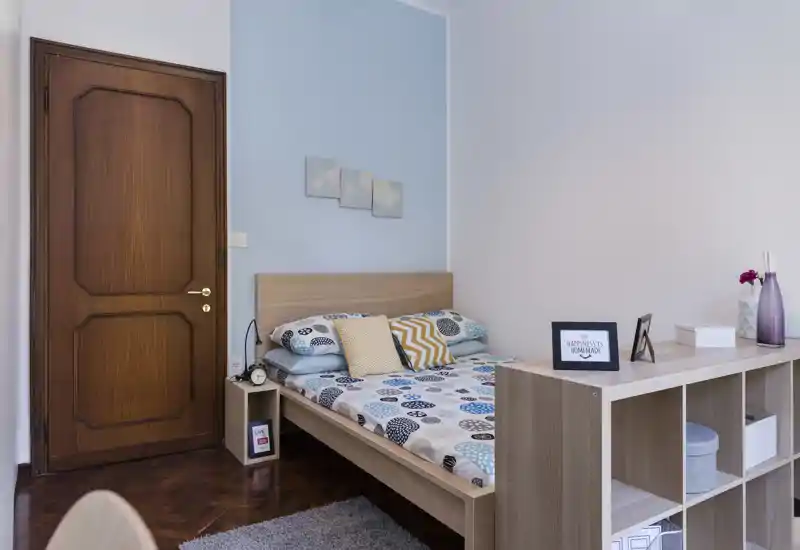 Bright, neatly styled bedroom with a double bed, patterned bedding and soft decorative cushions. Light wood furniture and a small shelving unit create a practical and welcoming layout ideal for short-term rentals.