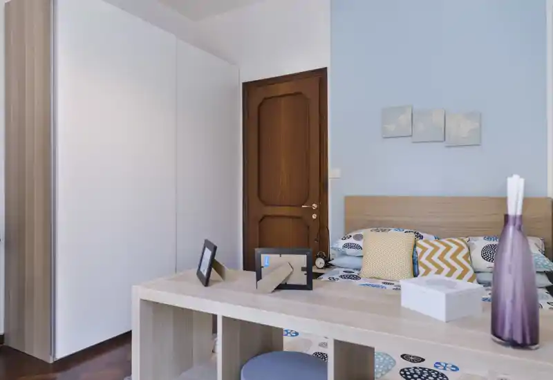 Bright, neatly styled bedroom with a wooden headboard, patterned bedding and decorative cushions. A desk/shelf unit sits at the foot of the bed and a wardrobe and wooden door are visible, giving a sense of practical storage and calm decor.