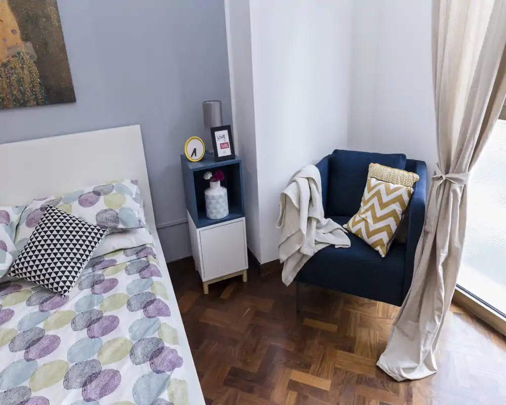 Camera da letto moderna e luminosa con copriletto a motivo, cuscini geometrici e una poltrona accogliente vicino alla finestra, perfetta per rilassarsi.