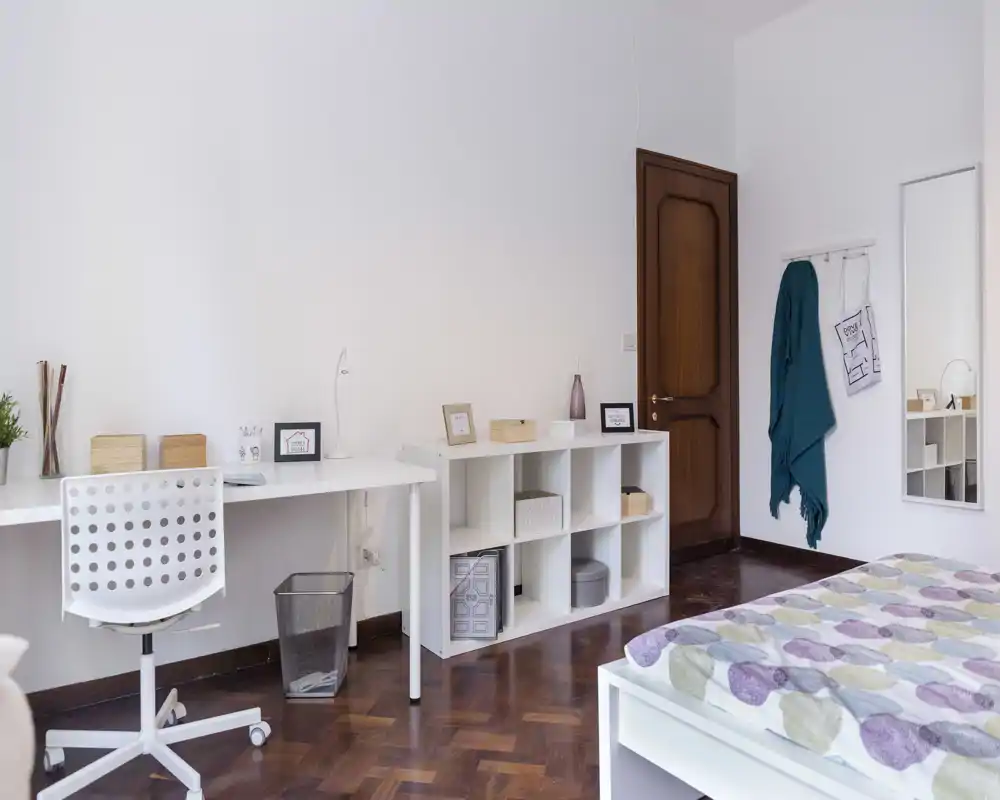 Camera da letto luminosa e ben allestita con struttura letto bianca e copriletto a fantasia, area studio con scrivania e sedia e scaffalatura ordinata — perfetta per un annuncio in affitto che punta su comfort e funzionalità.