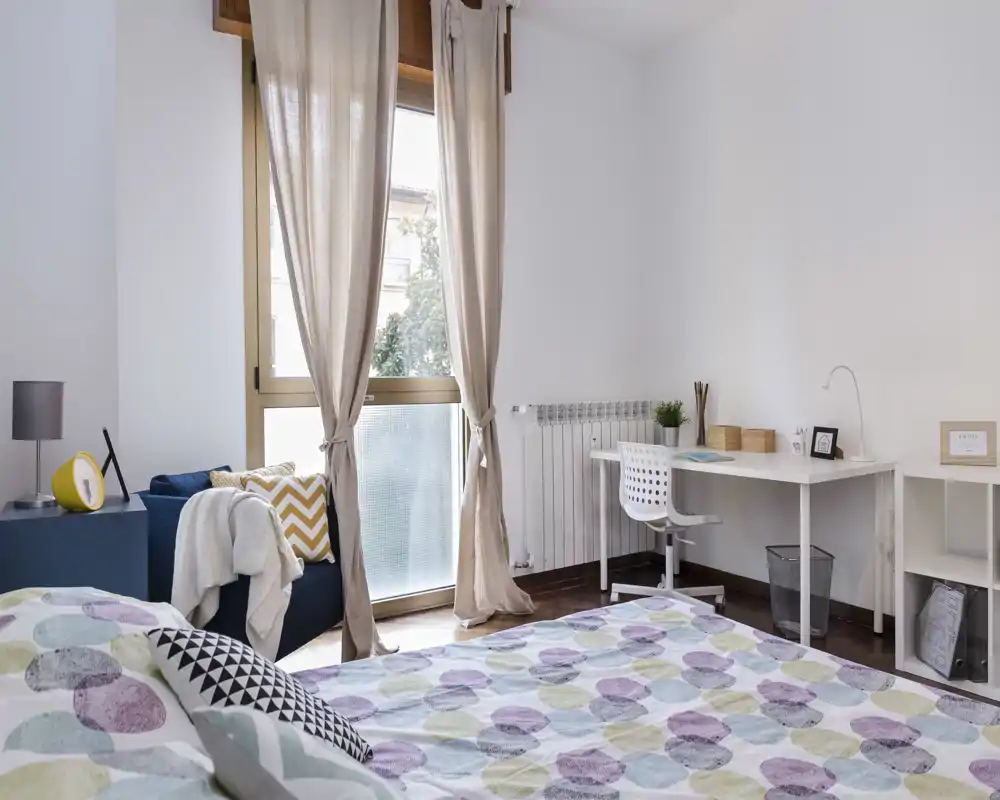 Camera da letto luminosa e ben arredata con copriletto a fantasia, ampia finestra con tende leggere che lasciano entrare la luce naturale, scrivania e comoda seduta — ideale per chi cerca una stanza tranquilla e soleggiata.