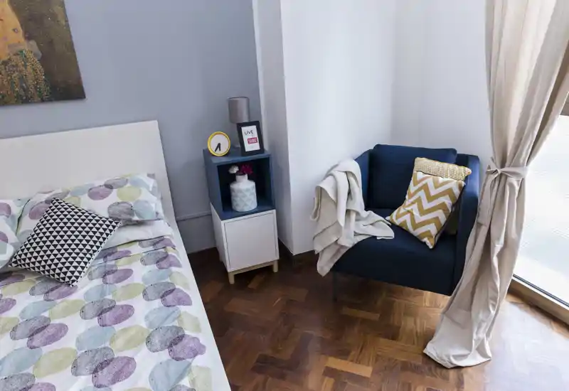 Camera da letto luminosa e arredata con letto a fantasia, comodino a cubi e una poltrona accogliente vicino alla finestra — ideale per annunci che valorizzano comfort e luce naturale.