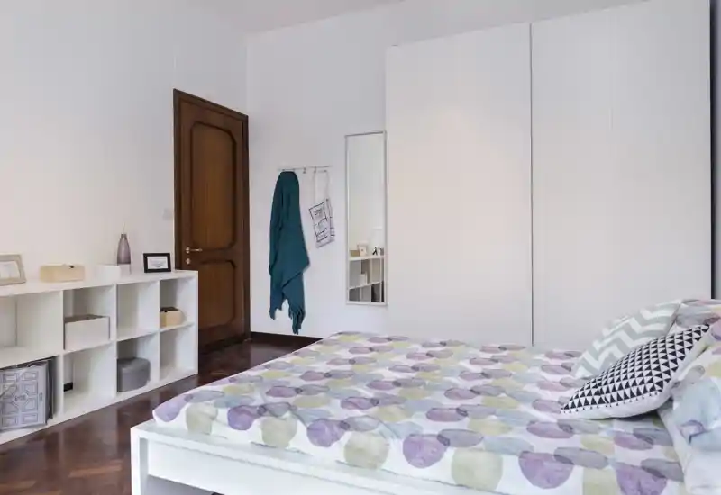 Camera da letto luminosa e ordinata con un ampio letto con copriletto fantasia, cuscini decorativi, grande armadio bianco e scaffalatura semplice—palette neutra e pavimento in legno lucido.