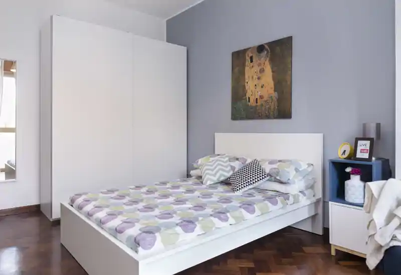 Camera da letto moderna e luminosa con letto bianco, biancheria a motivi e cuscini decorativi, ampia armadio bianco e quadro che crea un'atmosfera accogliente e fresca.