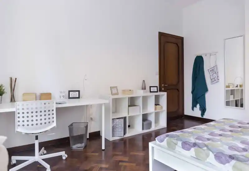 Camera da letto luminosa e ordinata con letto bianco e copripiumino a fantasia, scrivania e scaffalatura aperta — ideale per una singola o uno studente.