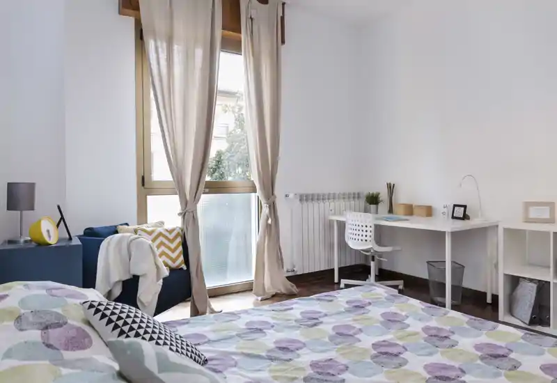 Camera da letto luminosa e ben arredata con copriletto a fantasia, ampia finestra con tende leggere che lasciano entrare la luce naturale, scrivania e comoda seduta — ideale per chi cerca una stanza tranquilla e soleggiata.