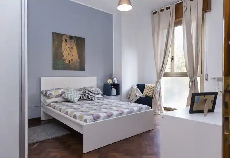 Camera da letto luminosa e ben arredata con letto bianco completo, biancheria a fantasia e cuscini decorativi. La luce naturale da una grande finestra e i toni neutri rendono l’ambiente accogliente e perfetto per gli annunci.