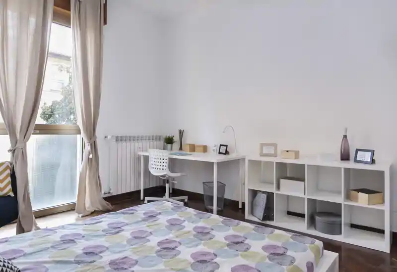 Camera da letto luminosa e minimal con copriletto a fantasia, scrivania e scaffalatura e una grande finestra che illumina naturalmente l'ambiente.