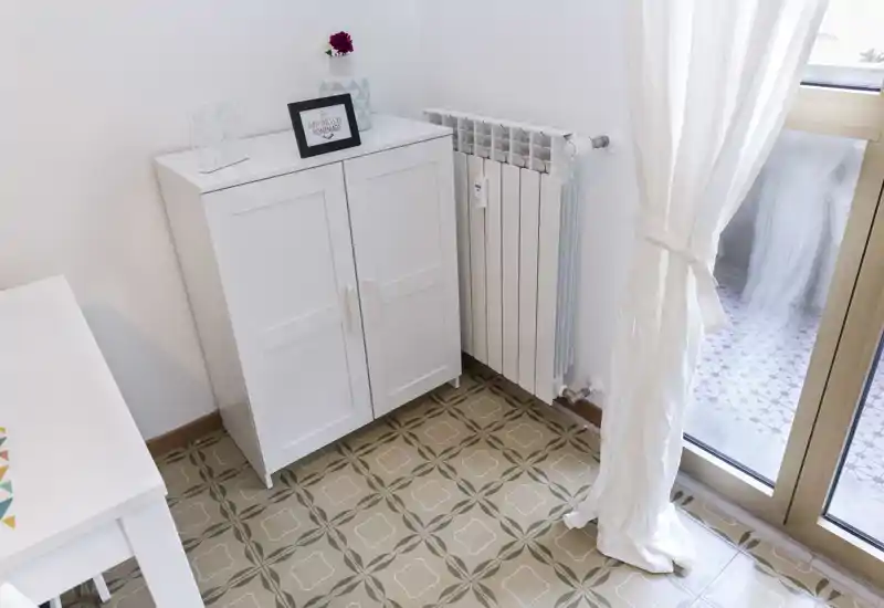Piccolo angolo luminoso di un soggiorno con un mobiletto bianco, termosifone e porta finestra sul balcone; pavimento piastrellato e arredo neutro creano un ambiente ordinato e arioso.
