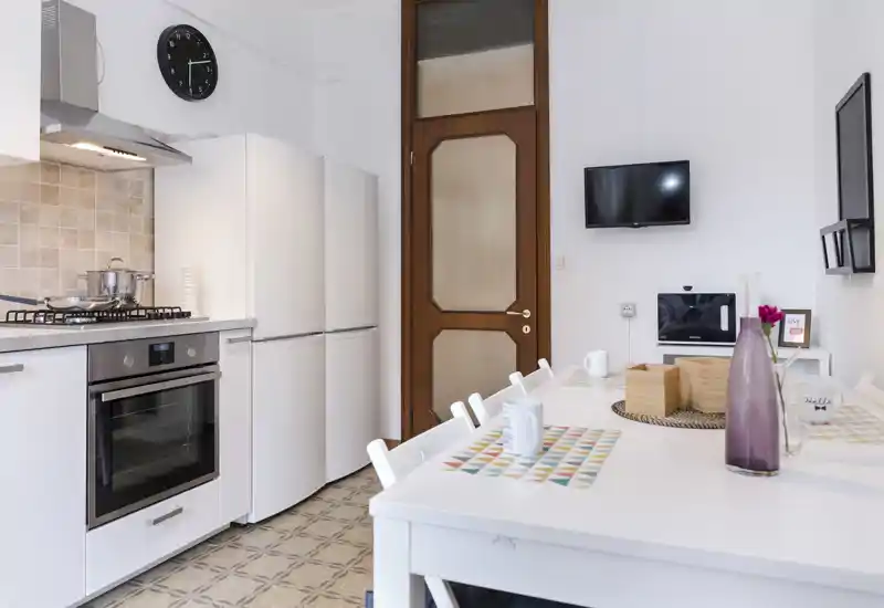 Cucina luminosa e moderna con mobili bianchi, forno incassato, frigorifero e un tavolo da pranzo apparecchiato; pulita e pronta all'uso.