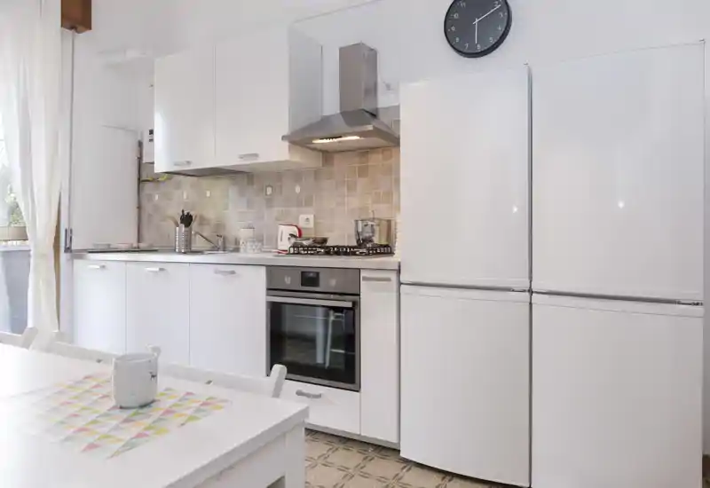 Cucina luminosa e moderna con mobili bianchi, forno incassato e piano cottura a gas, grande frigorifero e piccolo angolo pranzo vicino alla finestra.