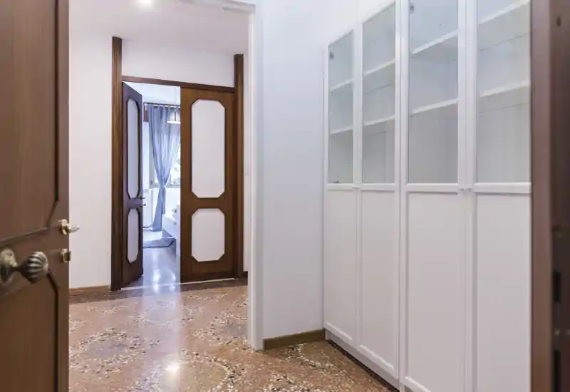 Ingresso luminoso e ordinato con armadietti bianchi a muro e pavimento in terrazzo decorato che conduce a doppie porte in legno e a una vista sull'ambiente adiacente.