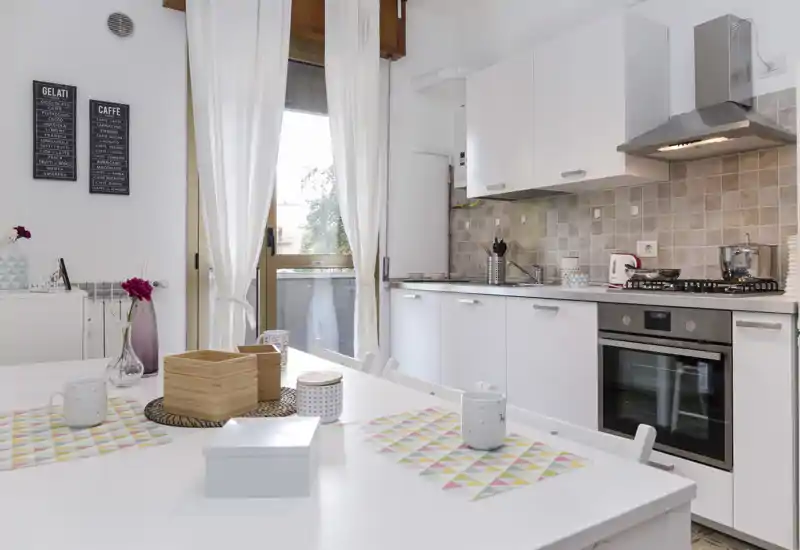 Cucina moderna e luminosa con mobili bianchi, forno incassato e tavolo da pranzo con tovagliette colorate. La luce naturale dalla finestra rende l'ambiente accogliente e ben arredato, perfetto per cucinare e pranzi informali.