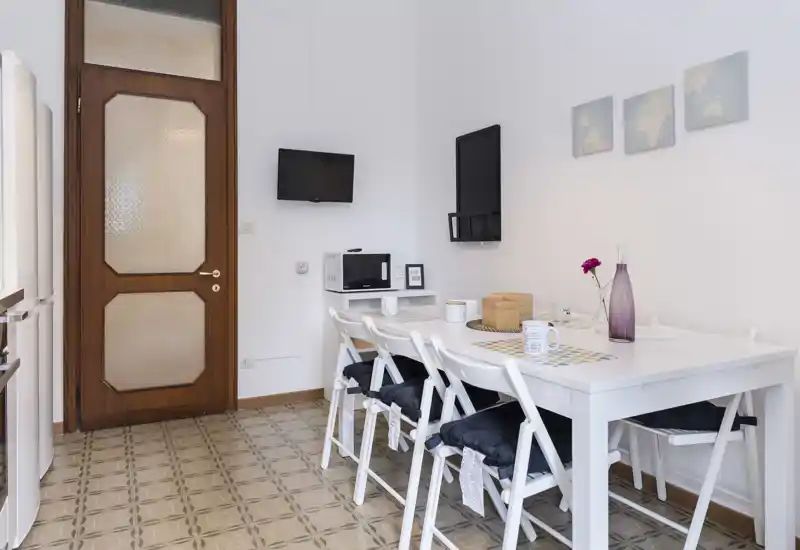 Zona pranzo luminosa e ordinata con tavolo bianco per più persone, sedie pieghevoli con cuscini e semplici quadretti a parete — perfetta per pasti in compagnia.