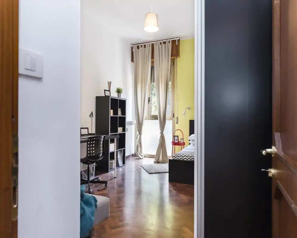 Camera da letto luminosa e accogliente con pavimento in parquet, letto singolo, scrivania e scaffali—ideale per studenti o professionisti che cercano uno spazio funzionale e ben illuminato.
