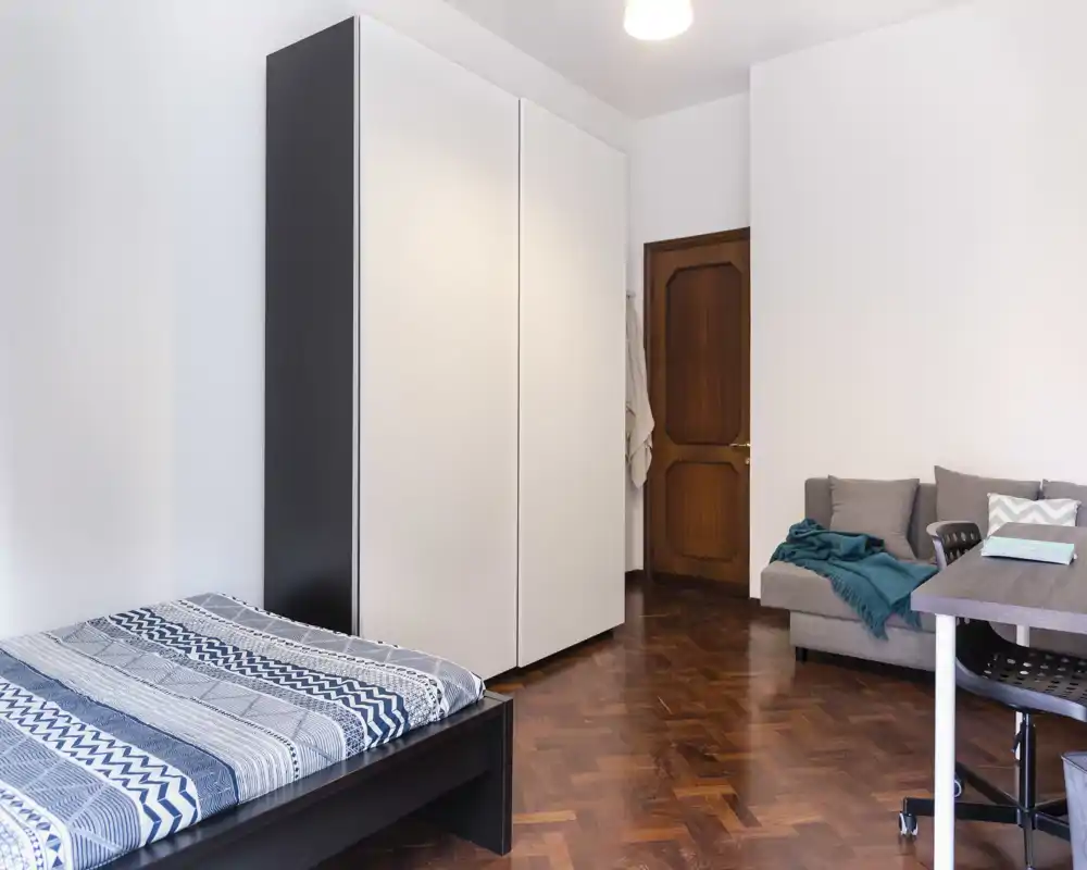 Camera da letto luminosa e arredata con semplicità: letto matrimoniale in primo piano, ampia armadiatura bianca e piccolo angolo con divano/tavolo; pavimento in parquet e pareti neutre per un ambiente pulito e accogliente.