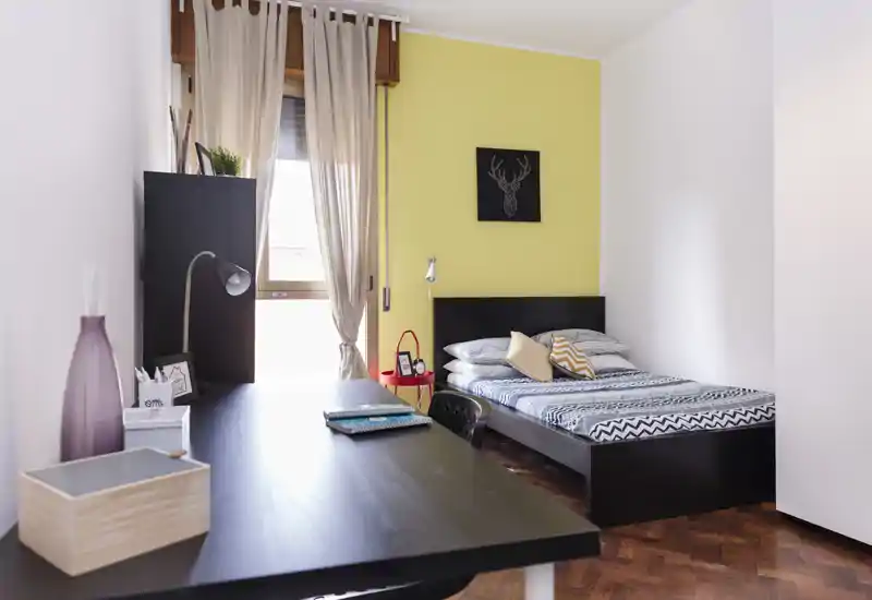 Camera da letto luminosa con letto matrimoniale, biancheria a fantasia e un'area studio in primo piano. La stanza ha una parete gialla calda, pavimento in parquet e tende leggere che filtrano la luce naturale.