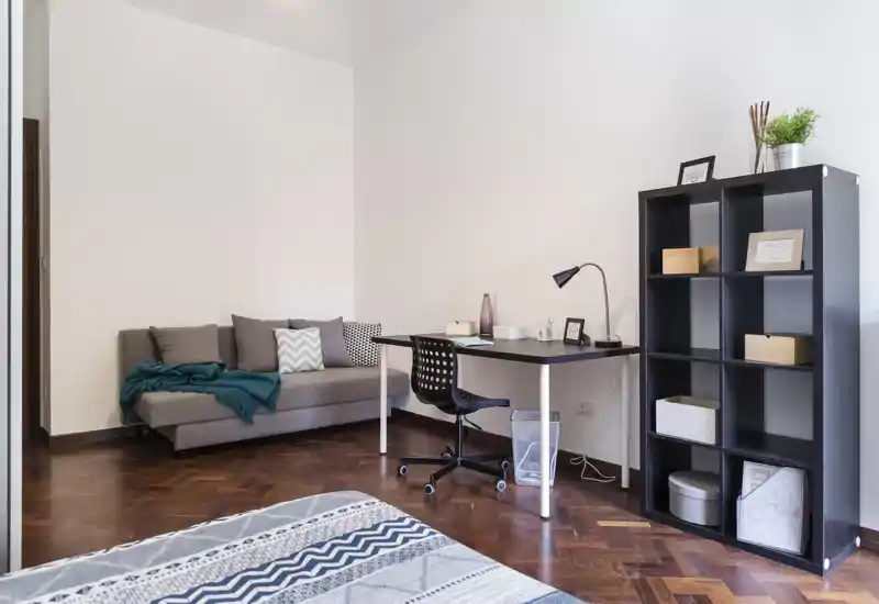 Camera da letto luminosa e ben arredata con letto in primo piano, un piccolo divano e una scrivania ordinata — ideale per affitti brevi o studenti.