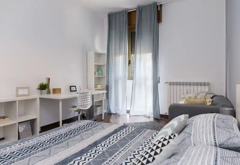 Camera da letto luminosa e moderna con biancheria a motivo, una scrivania lungo la parete e tende leggere che lasciano filtrare la luce naturale. La disposizione include un letto confortevole in primo piano e un piccolo divano vicino alla finestra, per uno spazio funzionale e arioso.