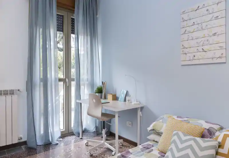 Bright, pastel bedroom with a small desk by the balcony. The bed with patterned cushions and soft textiles sits in the foreground and a study corner with natural light creates a welcoming atmosphere.