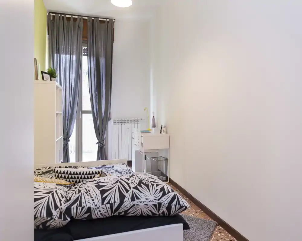 Chambre lumineuse et soignée avec housse de couette à motif, rideaux légers et petit bureau près de la fenêtre — idéale pour une location confortable.