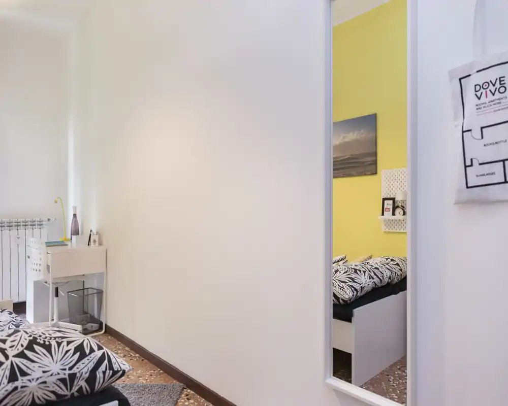 Coin de chambre moderne et lumineux avec couette à motifs, petit bureau et miroir reflétant le lit — espace chaleureux et bien éclairé, parfait pour une annonce locative.