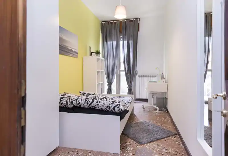Chambre moderne et lumineuse avec couette à motifs, étagère et voilages laissant passer la lumière naturelle — confortable et bien mise en scène.