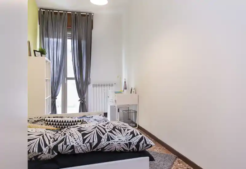 Chambre lumineuse et soignée avec housse de couette à motif, rideaux légers et petit bureau près de la fenêtre — idéale pour une location confortable.