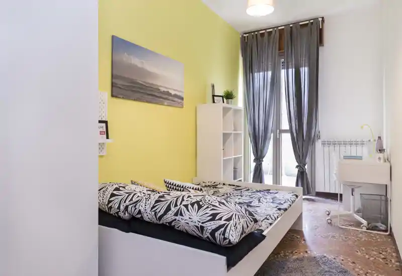 Chambre moderne et lumineuse avec couette à motifs, mur jaune accentué et grande fenêtre donnant sur le balcon offrant une belle lumière naturelle. Bureau compact et étagères pour le rangement.