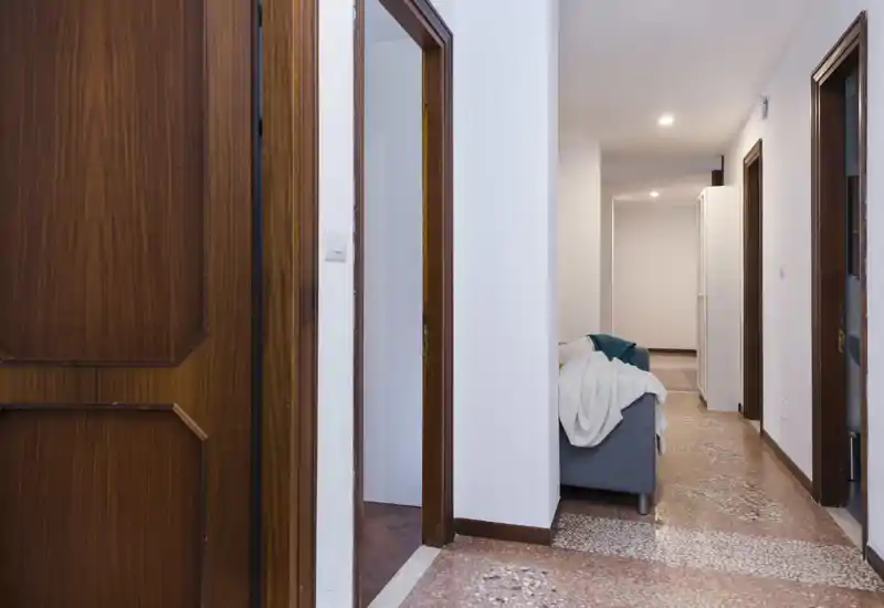 A hallway view showing a small sitting area with a sofa partially visible; white walls, wood door frames and terrazzo floor create a simple, neutral interior.