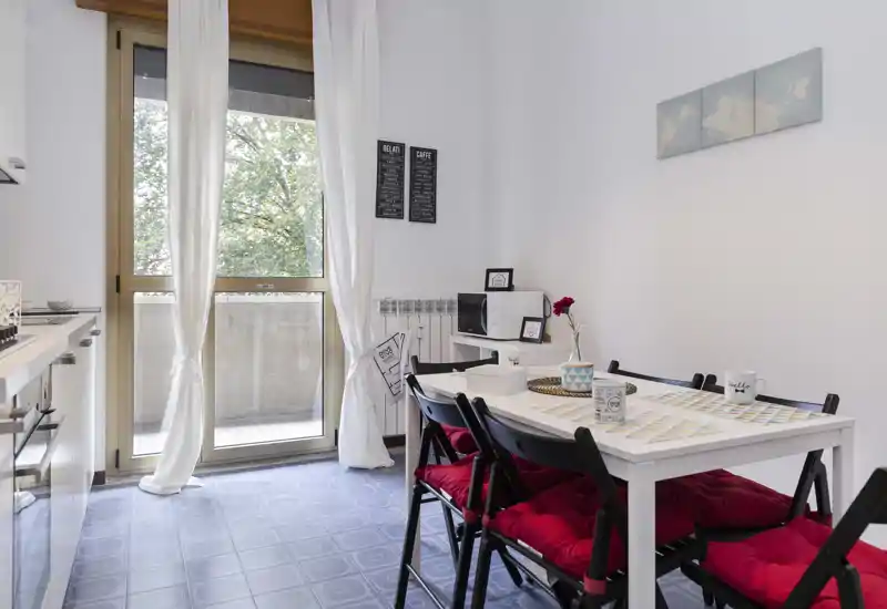 Bright, modern kitchenette with a dining table for four, red seat cushions, and a large glass door opening to a small balcony that fills the room with natural light.