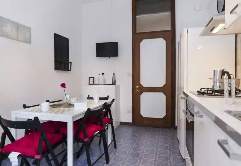 Tidy dining area integrated with a compact kitchen: white table with cushioned folding chairs, simple table setting and nearby kitchen counter with stove — ideal for cozy meals in a small apartment.