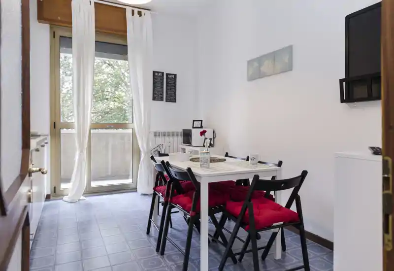 Bright, cozy dining area with a white table and four black folding chairs with red cushions, next to a balcony door that brings natural light.