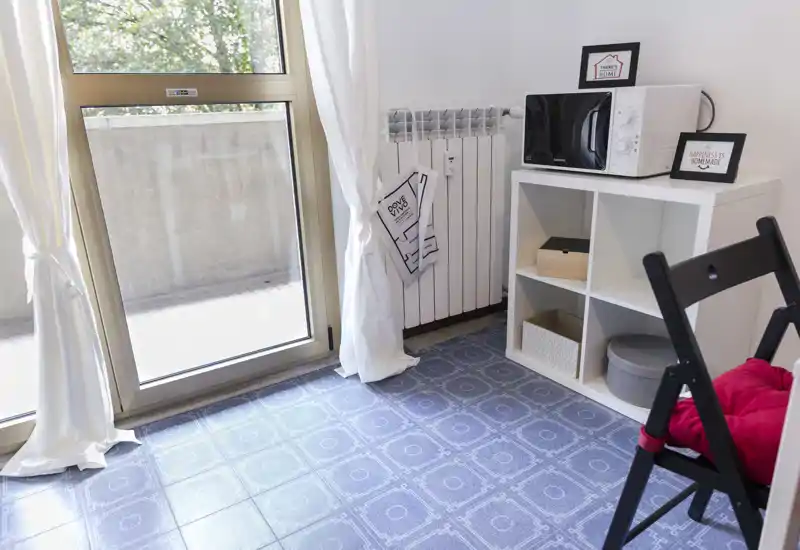 Small living area with a large glass door letting in abundant natural light, simple shelving with a microwave and a folding chair with a cushion.