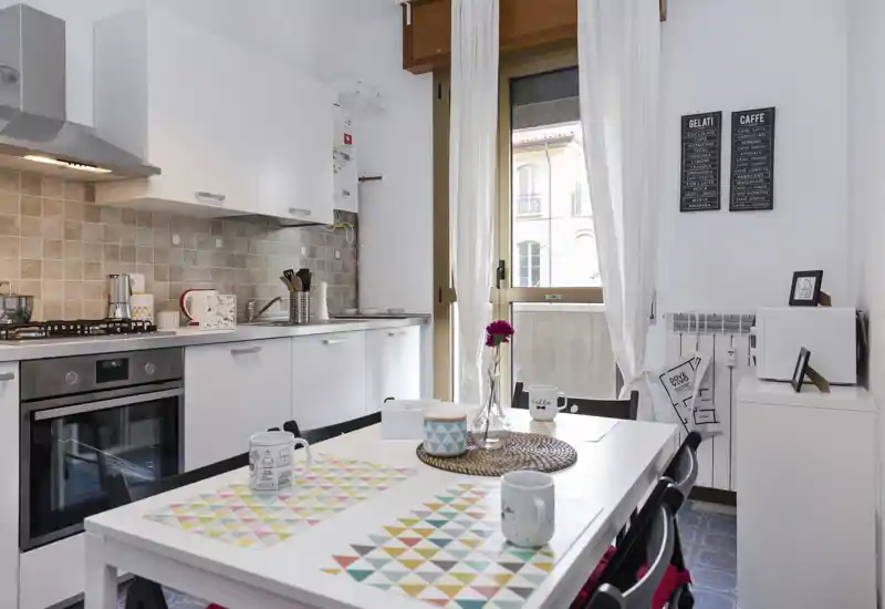 Bright modern kitchen with white cabinets, built-in oven and a small dining table set with colorful placemats and mugs; a window/door brings natural light and creates a welcoming atmosphere.