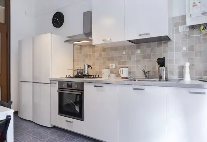 Modern, compact kitchen with white cabinets, built-in oven, gas hob and a double-door fridge. Neutral tiled backsplash and small accessories create a tidy, functional space.