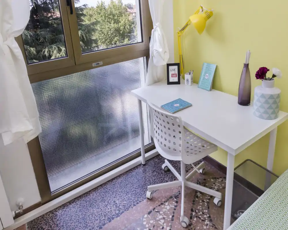 Small bedroom corner with a white desk and chair beside a large window; a portion of the bed is visible at the right edge, creating a bright, functional study nook.