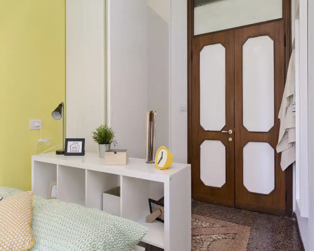 Bright, modern bedroom corner with patterned bedding, a low shelving headboard, decorative cushions and a wooden door — neat styling and pleasant natural light.