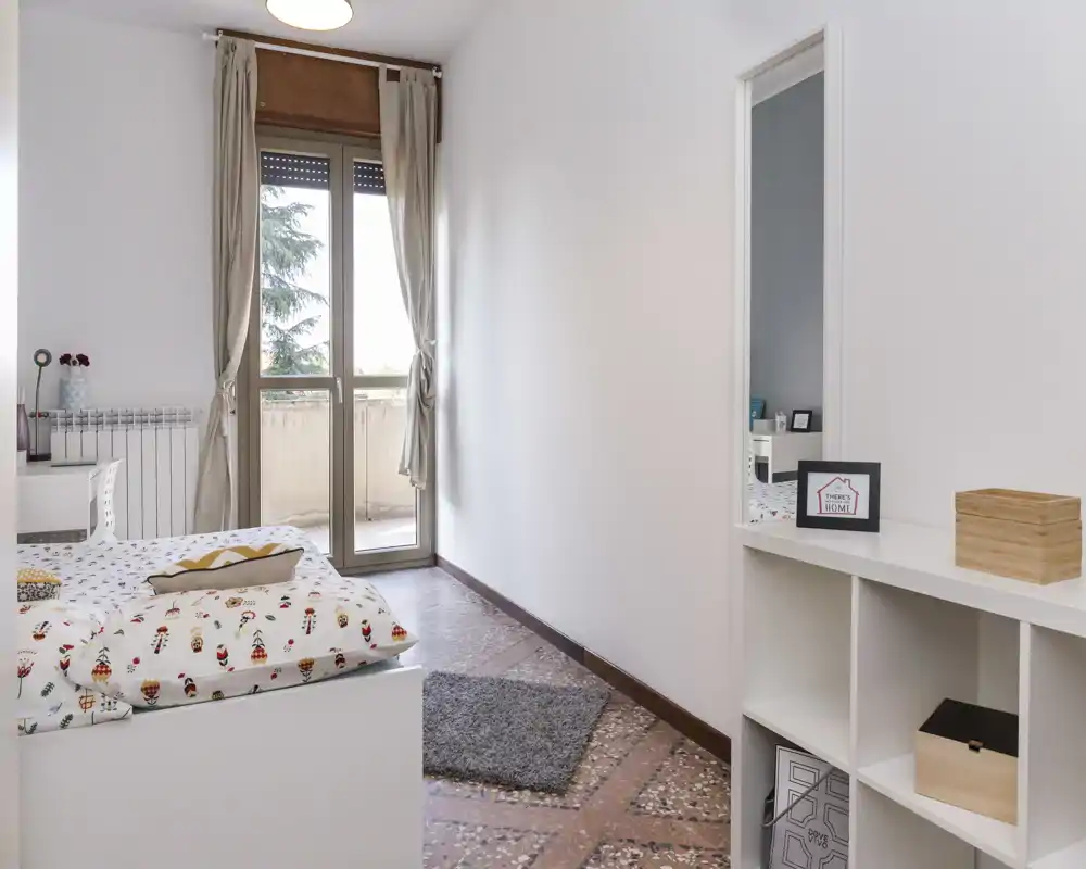 Camera da letto luminosa e moderna con biancheria a fantasia, porta balcone che illumina l'ambiente e semplici scaffali bianchi per tenere tutto in ordine.