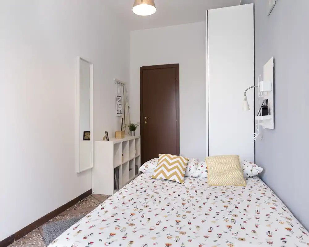 Camera da letto doppia luminosa e ben allestita con biancheria a fantasia, cuscini decorativi e un armadio alto. La stanza è ordinata e accogliente.