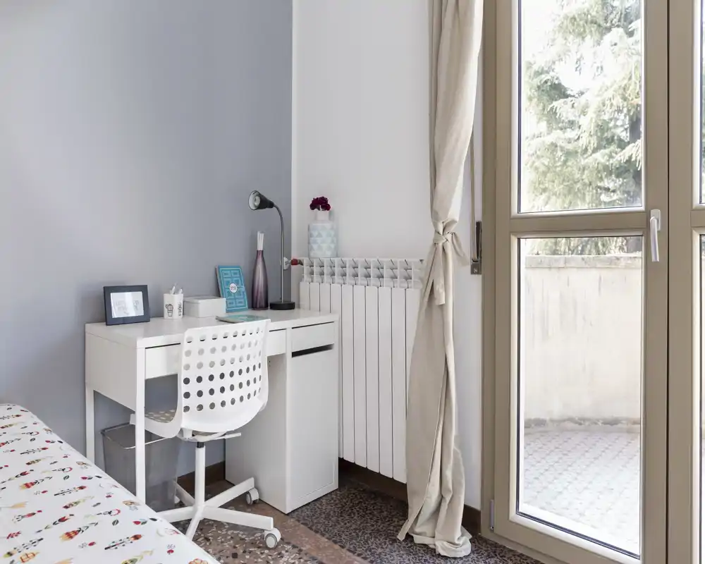Angolo di camera luminoso e moderno con copriletto a fantasia, scrivania bianca compatta e sedia ergonomica vicino a una grande finestra che lascia entrare luce naturale.