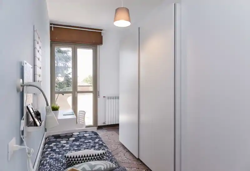 Camera singola compatta con letto ben rifatto, scrivania vicino a una grande finestra e armadio bianco alto; spazio luminoso, moderno e ordinato, ideale per uno studente o una persona sola.
