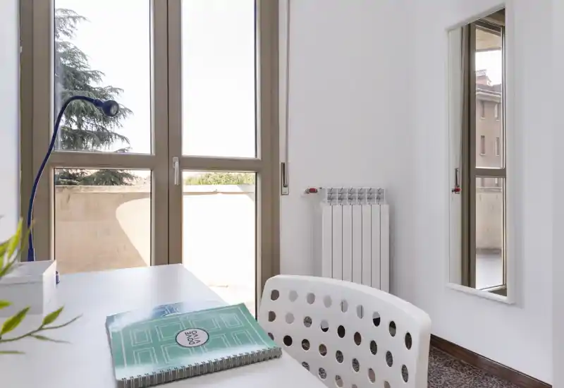 Stanza luminosa e ordinata con una scrivania vicino a grandi finestre che lasciano entrare molta luce naturale; una sedia moderna forata, una lampada da tavolo e una piccola pianta creano un angolo di lavoro piacevole.