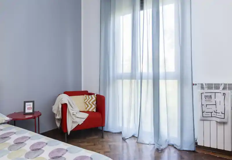 Bright, cozy bedroom corner with patterned bed linens, a red accent armchair and soft natural light from large windows—ideal for restful stays.