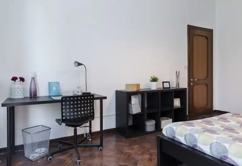 Bright, tidy bedroom with a patterned duvet, a black platform bed visible at the right and a workspace with desk and chair at left. Neutral walls, hardwood floors and simple shelving make this room feel modern and functional.