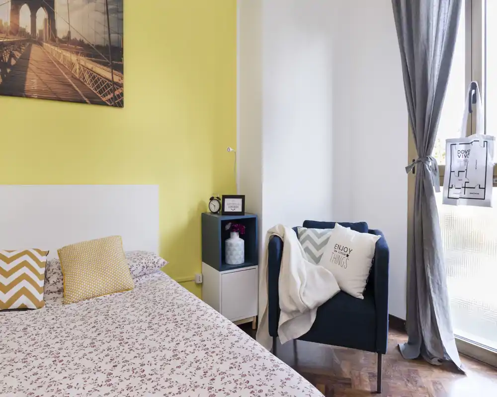 Camera da letto luminosa e ben arredata con letto decorato, cuscini coordinati e una poltrona accogliente accanto alla finestra.